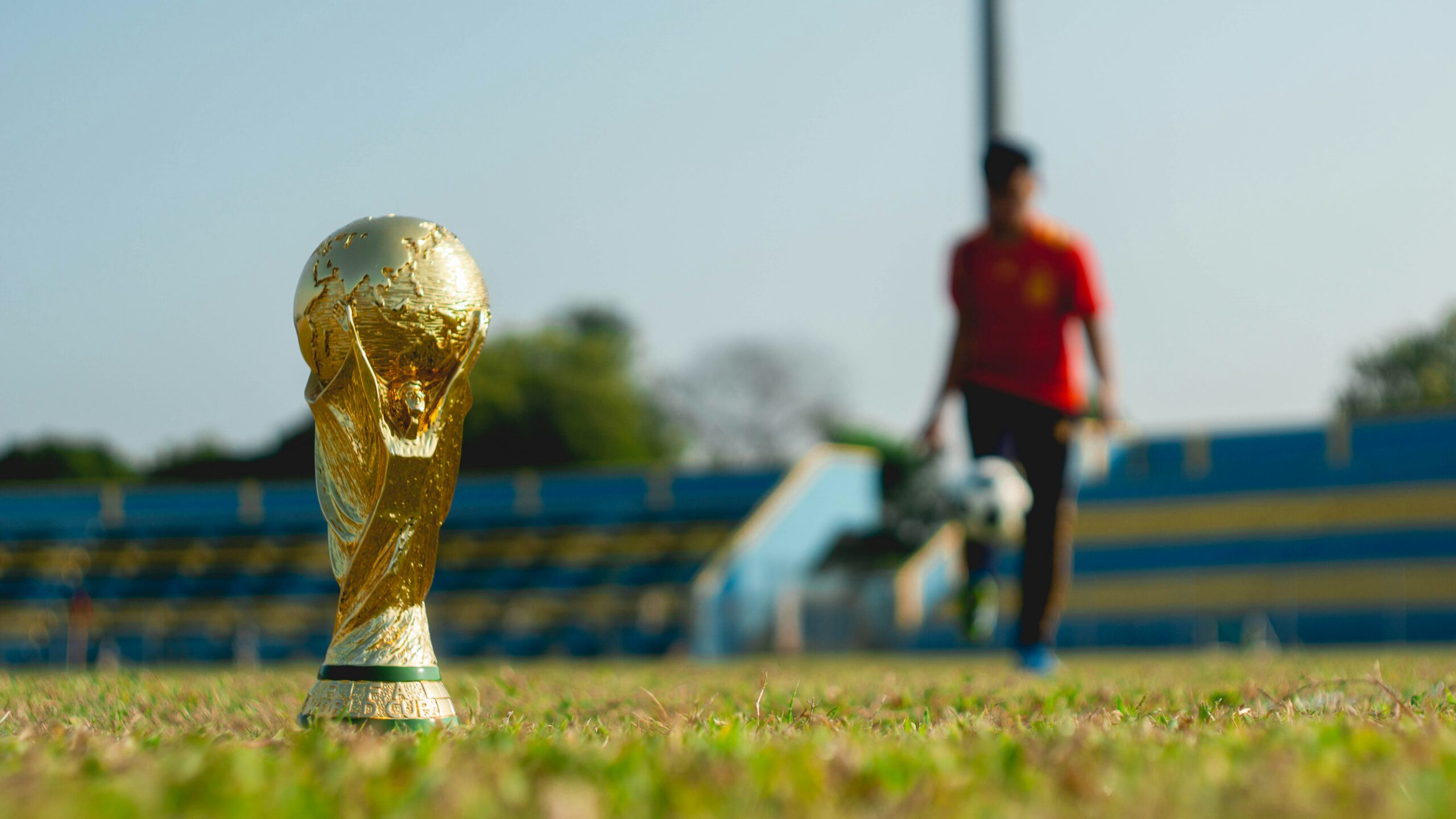 In focus, foreground: the World Cup trophy sitting on the grass of a football pitch. In the background, out of focus, a football player kicks a ball.